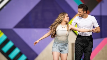 Karol Sevilla y un hombre joven, ambos sonrientes, mirándose y tomados de la mano, posan frente a una pared con patrones geométricos de colores púrpura, azul verdoso y amarillo