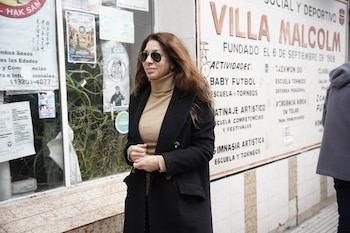 Mujer con cabello castaño, gafas de sol, suéter cuello tortuga beige y abrigo negro. De pie frente a una vidriera y un cartel blanco en la calle