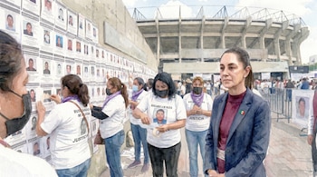 Pintura acuarela: Mujeres pegan carteles de 'DESAPARECIDO' en muro. Claudia Sheinbaum en primer plano con pin bandera mexicana. Detrás, estructura de estadio y multitudes.