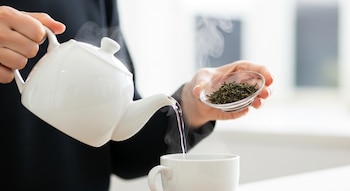 Primer plano de manos vertiendo agua hirviendo de una tetera blanca en una taza, mientras se sostiene un cuenco de cristal con hojas de té verde secas.
