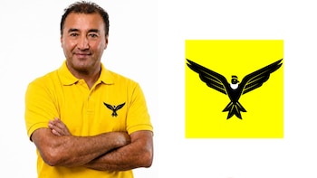 Un hombre con polo amarillo y los brazos cruzados posa al lado del logotipo de un ave negra sobre fondo amarillo