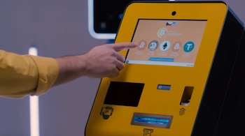 Mano de persona tocando pantalla de cajero automático amarillo con logotipos de Bitcoin, Dogecoin, Ethereum, Tether y opciones de compra. Incluye ranura de billetes, escáner QR y lector de huellas