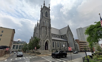 Fachada de una iglesia de piedra gris con una alta torre central en una esquina de calle con vehículos estacionados y señales de tránsito