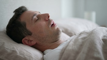 Un hombre duerme boca arriba en una cama con una almohada clara, su boca está abierta y sus ojos cerrados, en un primer plano lateral que muestra detalles faciales.