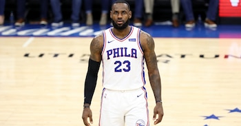 Jugador de baloncesto con barba, vistiendo camiseta blanca con 'PHILA 23', manga negra y pulsera negra, de pie en una cancha de madera