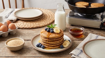 Hot cakes con arándanos, plátano, menta y miel en un plato sobre mesa de madera. También hay huevos, leche, un bol de granos de amaranto y una sartén.