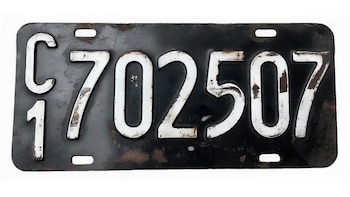 Chapa patente de automóvil rectangular negra con letras y números blancos desgastados, C 702507, y el número uno debajo de la letra C