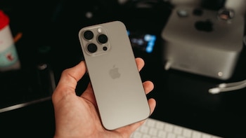 Una mano sostiene un teléfono inteligente iPhone plateado con tres cámaras traseras sobre un escritorio con teclado y ordenador
