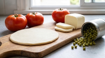 Vista de cerca de una tabla de madera con un disco de masa, tomates rojos, un bloque de muzzarella cortada y una lata de arvejas volcadas.