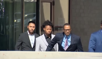 Un joven con traje gris y cabello rizado, Karmelo Anthony, camina flanqueado por dos hombres vestidos de traje fuera de un edificio