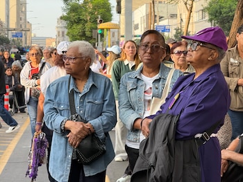 Tres mujeres de pie en una calle urbana; una viste hábito y gorra morados, mientras otras personas permanecen detrás