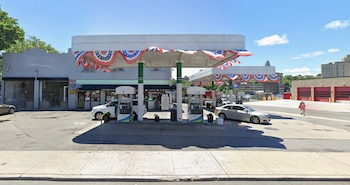 Una estación de servicio con cuatro surtidores de combustible, dos vehículos, un local comercial con banderas, una calle y edificios