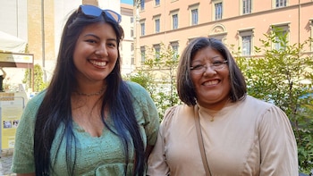 Dos mujeres sonrientes de pie al aire libre, con cabello oscuro, una con gafas y la otra con gafas de sol en la cabeza. Un edificio rosado y árboles de fondo