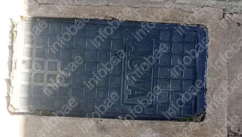 Primer plano de una tapa de alcantarilla rectangular oscura con patrón de rejilla y la palabra 'AGUA' grabada, incrustada en cemento agrietado