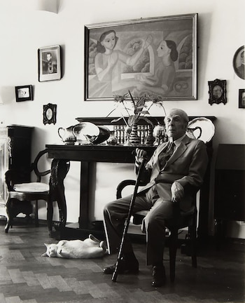 Fotografía en blanco y negro de Jorge Luis Borges, un hombre mayor de traje, sentado con un bastón. Un gato blanco se recuesta en el suelo de parqué, bajo una mesa de madera con objetos y un cuadro grande