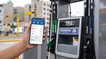 Mano que sostiene un teléfono móvil con una aplicación de precios de combustible frente a un surtidor en una estación de servicio.