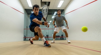 Dos hombres juegan squash en una cancha interior. Uno, de camiseta azul, golpea una pelota amarilla con una raqueta. El otro, de camiseta gris, observa detrás.