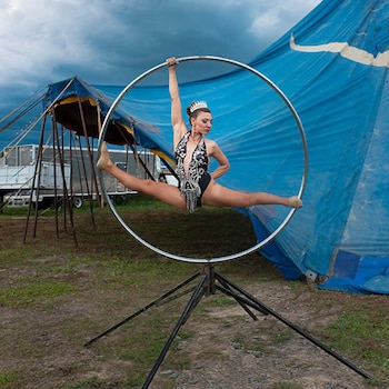Una mujer realiza una apertura de piernas en un aro metálico montado sobre una base metálica, frente a una carpa de circo azul