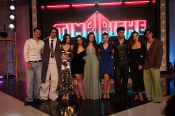 Nueve jóvenes vestidos de gala posan alineados en un escenario frente a un letrero luminoso que dice Timbiriche