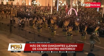 Desfile nocturno con danzantes de trajes amarillos y azules con penachos, una banda de música, barreras de seguridad y una multitud de espectadores