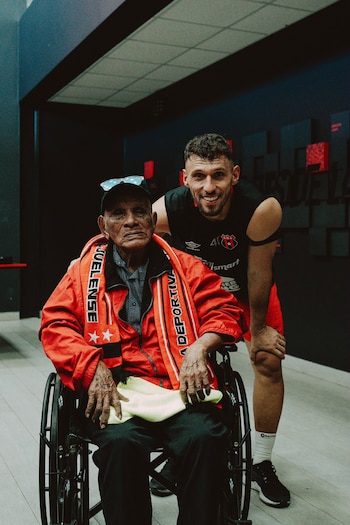 Hombre joven de pie junto a hombre mayor en silla de ruedas, ambos sonrientes. El hombre joven usa camiseta negra, el mayor chaqueta roja, gorra y bufanda