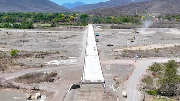 Vista aérea de uma ponte de concreto em construção sobre terras rochosas e estéreis, com ladrões e montanhas no horizonte sob um céu azul
