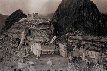 Fotografía monocromática de Machu Picchu mostrando terrazas agrícolas y estructuras de piedra incas parcialmente cubiertas por vegetación, con montañas al fondo