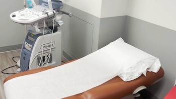 Máquina de ultrasonido Voluson junto a una camilla de examen con una cubierta desechable blanca en una sala de paredes blancas y grises