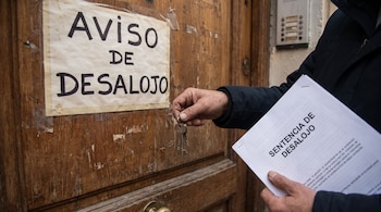 Una mano entrega llaves a otra que sostiene una sentencia de desalojo, frente a una puerta con un cartel de aviso de desalojo.