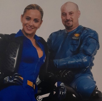 Una mujer y un hombre posan con trajes azules de motociclista, guantes y protecciones. El hombre usa una chaqueta con el logo de Noticieros Televisa