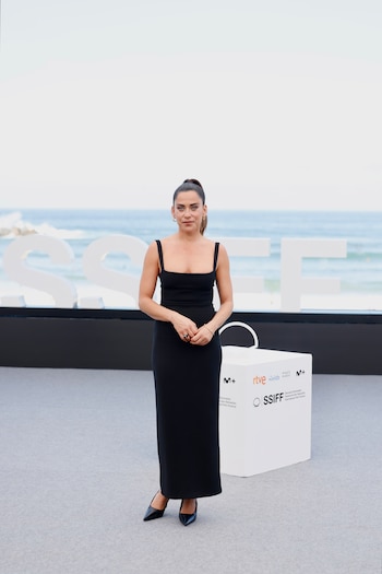 Una mujer con vestido negro y calzado de tacón posa frente a las letras del logotipo del festival y el mar al fondo