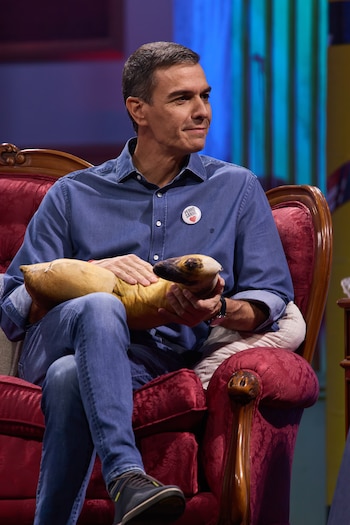 Pedro Sánchez lleva camisa azul con una chapa y vaqueros, sentado en un sillón rojo mientras sujeta un objeto alargado de tela