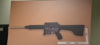 Un rifle de color negro con cañón metálico sobre una superficie plana marrón, junto a una regla de medición forense en forma de L