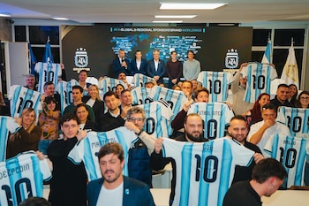 Un grupo de personas sostiene camisetas de fútbol argentinas celestes y blancas con el número diez. Cinco hombres están de pie al fondo frente a una pantalla de patrocinadores y banderas