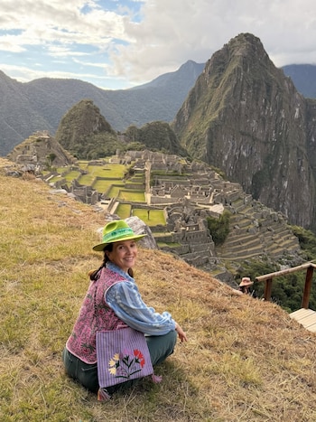 Mujer sonriente con sombrero verde, chaleco fucsia y bolso lila agachada sobre pasto seco, con las ruinas de Machu Picchu y montañas de fondo
