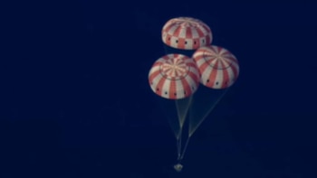 Tres paracaídas grandes con rayas rojas y blancas, anclados a una pequeña cápsula espacial, descienden contra un cielo azul oscuro