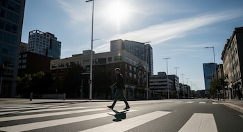 Hombre de perfil camina en un paso de cebra. Al fondo se ven edificios, postes de alumbrado público y un cielo azul con el sol brillando.