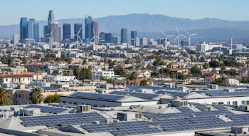 Vista aérea de Los Ángeles con tejados cubiertos de paneles solares, rascacielos modernos en el centro y turbinas eólicas al fondo con cielo azul claro.