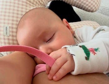 Primer plano de un bebé durmiendo, recostado sobre un hombro, con su pequeña mano agarrando una cinta rosa. Viste un body blanco con detalles de colores