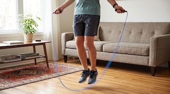 Hombre con camiseta, pantalones cortos y zapatillas deportivas salta una cuerda azul en el interior de una casa, con un sofá y una planta visible.