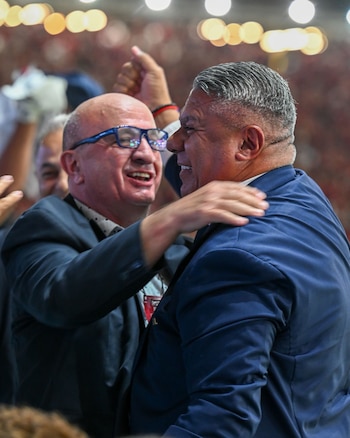 Dos hombres adultos se abrazan y sonríen en primer plano. El de la izquierda lleva gafas azules, camisa estampada y traje. El de la derecha lleva traje oscuro y pelo canoso
