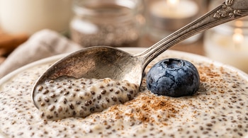 Cuchara metálica sobre un tazón de pudín de semillas de chía decorado con canela molida y un arándano azul fresco.