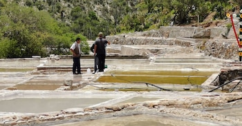 Tres hombres de pie entre terrazas escalonadas de piedra llenas de agua. Colinas verdes y cactus al fondo. Una cruz decorada a la derecha