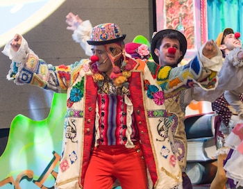 Osqui Guzmán y dos actores más vestidos de payasos con narices rojas, sombreros y chaquetas coloridas, en un escenario con utilería de libros