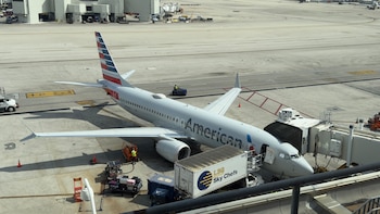Avión blanco de American Airlines siendo empujado por un tractor azul en la pista de un aeropuerto soleado, con un trabajador y edificios en el fondo