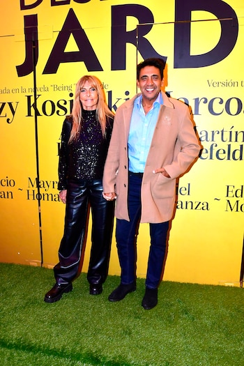 Dos personas, una mujer rubia y un hombre de cabello oscuro, posan sonrientes y de la mano sobre una alfombra verde que simula césped, frente a un fondo amarillo con texto