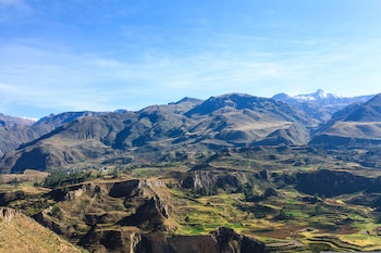 Vista panorámica de un valle montañoso con terrazas agrícolas verdes, laderas rocosas y un pico nevado al fondo bajo un cielo despejado