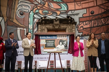 Cinco personas en un escenario aplauden mientras develan un billete de Lotería Nacional gigante con imágenes de educadores frente a un mural artístico