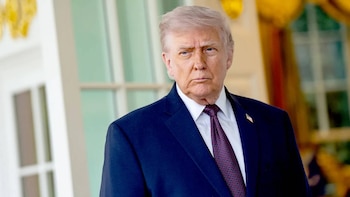 El Presidente de EE. UU., Donald Trump, con cabello gris, traje azul, camisa blanca y corbata roja, de pie con una expresión seria frente a un fondo borroso