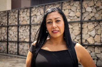 Retrato de una mujer de cabello negro largo y liso, mirada al frente, labios pintados de rojo. Viste una blusa negra. Fondo de pared con piedras